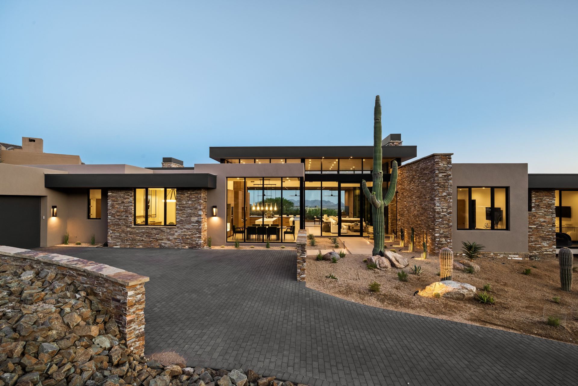 Modern single-story desert home at dusk with stone walls, lit floor-to-ceiling windows, a paver driveway and a saguaro cactus